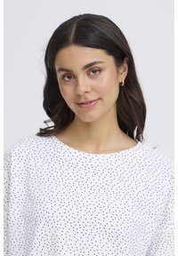 Mladá žena s tmavými vlasmi, ktorá nosí zlaté kruhové náušnice a bielu blúzku s malými čiernymi bodkami, jemne sa usmieva na svetlom pozadí.