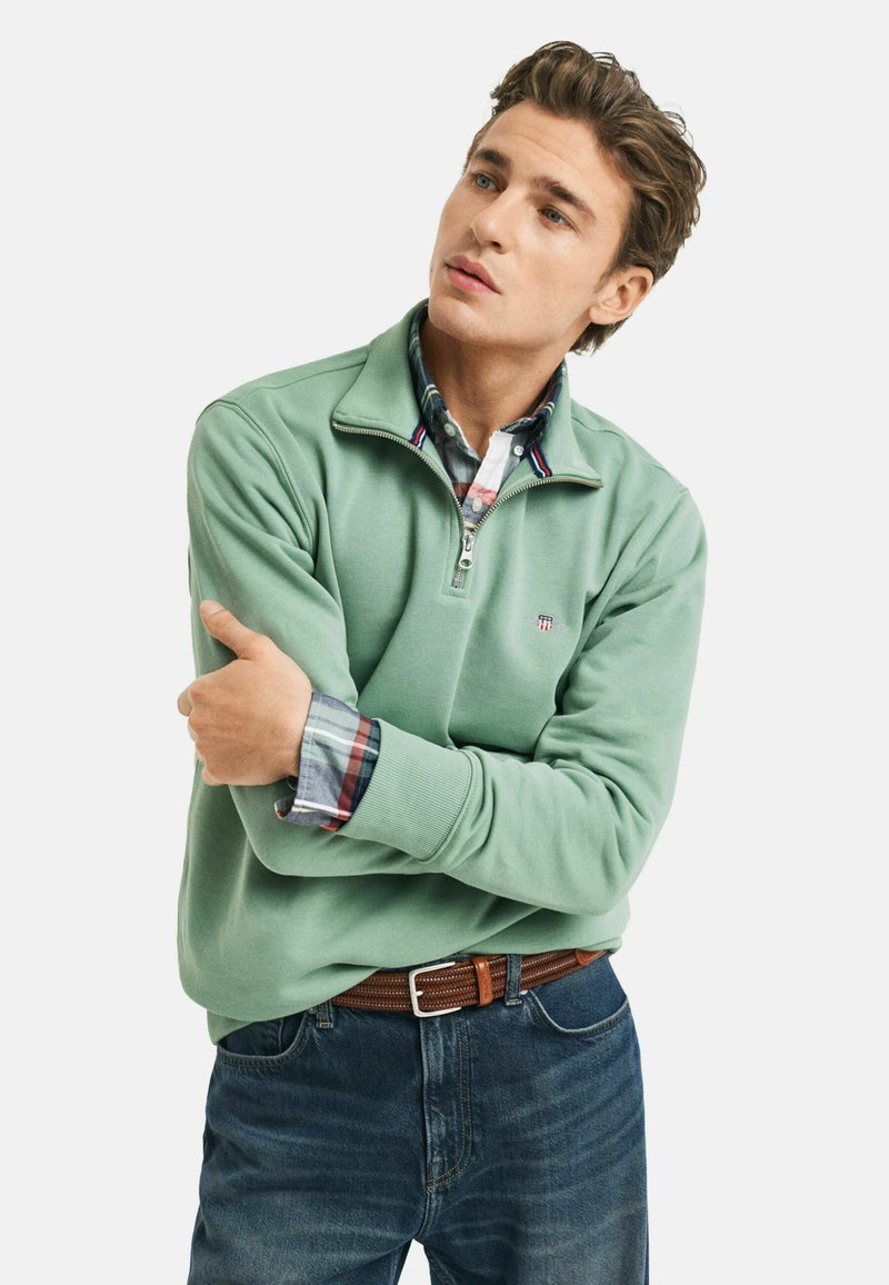Grüner Reißverschluss-Pullover mit weicher Textur, über einem karierte Hemd getragen. Auffällige Details sind ein kleines Logo und ein kontrastierender Kragen. Kombiniert mit Jeans.