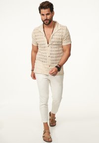Beige gestricktes kurzärmeliges Hemd mit Rautenmuster, vorne geknöpft, kombiniert mit weißen Strukturhosen und hellbraunen Sandalen.