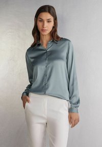 Camisa de satén en un tono verde azulado apagado, con cuello, mangas largas y una textura suave, combinada con pantalones blancos a medida.