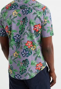 Kortärmad skjorta i ljuslila tyg, prydd med ett blommönster som innehåller röda, gula och blå blommor samt gröna blad. Strukturerad yta.