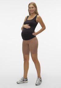 Svart tank topp, lysebrune høyt skårne shorts og grå sportsko; modellen poserer med hendene på magen, kort hår, nøytral bakgrunn.