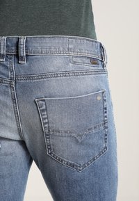 Světle modré džínové kalhoty s zakřiveným designem zadní kapsy, obsahující malé kovové logo a mírně vybledlou texturu.