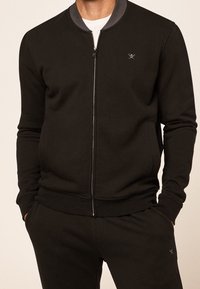 Sudadera negra con cremallera y cuello gris, con puños y dobladillo acanalados. Presenta un sutil logo en el pecho y bolsillos laterales. Textura de tela suave.