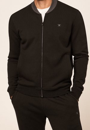 Felpa nera con zip e colletto grigio, polsini e orlo a costine. Presenta un logo discreto sul petto e tasche laterali. Tessuto con una consistenza liscia.