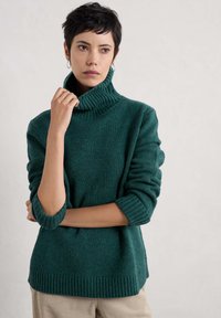 Teal stickad tröja med hög krage och ribbade mudd, med en avslappnad passform och textilstruktur. Subtila variationer i färg genom hela plagget.