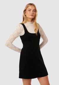 Czarna sukienka z korduroy o szerokich ramiączkach, w sylwetce A-linii, noszona na białej koronkowej bluzce z długim rękawem. Gładka tekstura, minimalistyczny design.