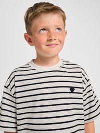 Młody chłopiec z krótkimi jasnobrązowymi włosami, ubrany w biało-czarną koszulkę w paski, patrzy w górę i lekko się uśmiecha na jednolitym tle.