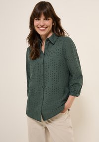 Chemise verte à manches trois-quarts, dotée de détails perforés et d'un col structuré. Tissu léger avec une texture lisse.