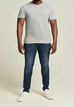 Homme debout portant un t-shirt à manches courtes gris clair uni, un jean bleu foncé et des baskets blanches sur un fond neutre.