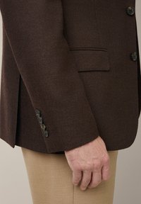 Brązowa teksturowana marynarka z zapięciem na guzik i pojedynczą kieszenią na piersi, połączona z jasnobeżowymi spodniami. Klasyczny design z dopasowanym krojem.