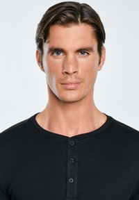 Jaunas vyras su trumpais rudais plaukais ir mėlynomis akimis, vilkintis juodą ribuotą Henlio marškinius, žiūrintis tiesiai priešais, stovintis priešais paprastą šviesią foną.