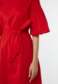 Rotes Kleid mit gepufften kurzen Ärmeln und einem gebundenen Taillengürtel, dargestellt an einer Person von der Schulter bis zur Mitte des Oberschenkels vor einem weißen Hintergrund.