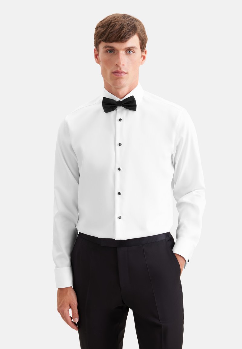 Homme portant une chemise blanche avec des boutons noirs, un nœud papillon noir et un pantalon de costume noir sur un fond clair uni.