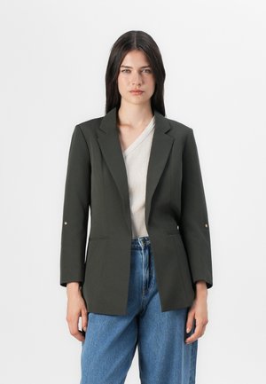 Model draagt een donkergroene op maat gemaakte blazer over een lichte v-hals top, gecombineerd met hooggetterpte blauwe jeans, staand tegen een effen achtergrond.