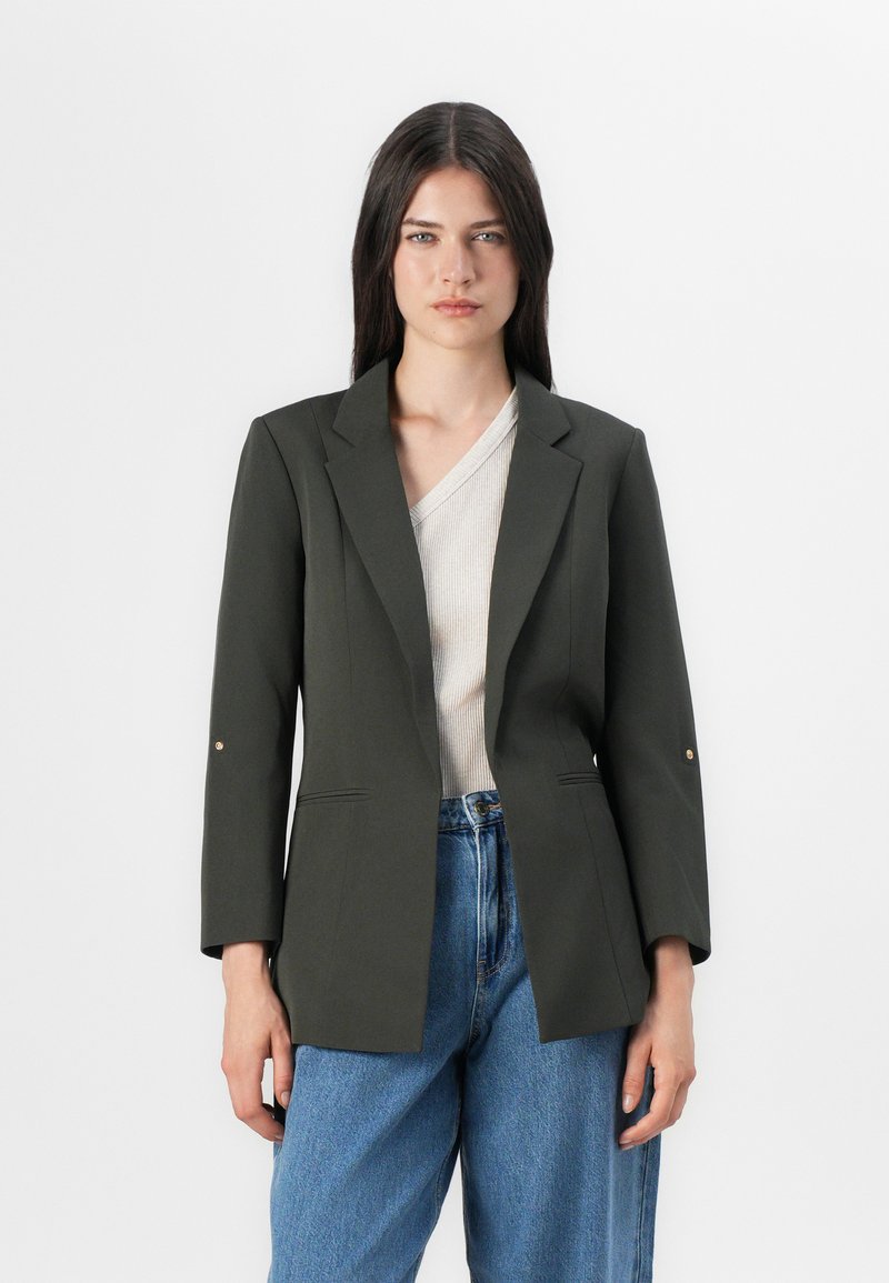 Modelo vistiendo un blazer entallado verde oscuro sobre una blusa ligera con escote en V, combinado con unos vaqueros de cintura alta azules, de pie frente a un fondo simple.