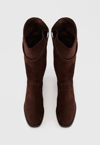 Stivali alti in suede marrone con una texture liscia, punta affusolata e dettagli curvi nella parte superiore. Bordo nero lungo la suola.
