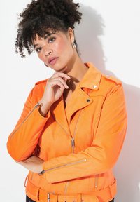 Veste de motard en daim orange avec poches zippées, taille ceinturée et accents en métal argenté. Elle présente un col à rabat et un tissu texturé.