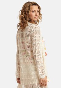 Leichtgewichtige, beige offene Strickjacke mit lässiger Passform, die ein strukturiertes Gewebe und lange Ärmel aufweist, getragen über einem bunten Blumenoberteil.