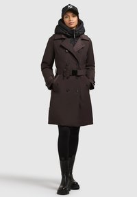 Tmavo hnedý trench coat s dvojitým zapínaním, opaskom a čiernymi detailmi; vrstvený s čiernou puffovou mikinou a tmavými legínami.