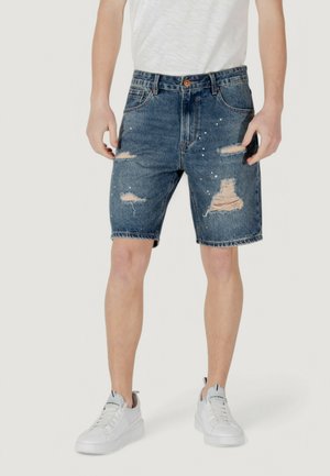 Shorts in denim con una lavatura blu, dettagli strappati e orli frangiati. Indossati con una t-shirt grigia chiara e sneakers bianche.