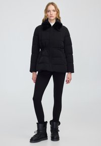 Zwarte pufferjack met een bontkraag, voorzien van een ritssluiting, elastische taille en zijzakken, gecombineerd met zwarte leggings en laarzen met bontvoering.