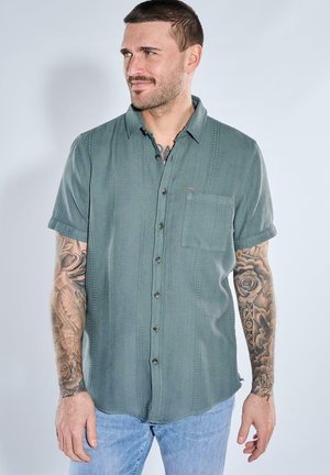 Mann mit kurzen braunen Haaren und Tätowierungen auf beiden Armen trägt ein grünes kurzärmeliges Hemd mit Knöpfen und hellblaue Jeans und schaut nach links.
