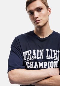 Mørkeblå atletisk t-shirt med korte ærmer, der har hvid tekst "TRÆN SOM EN MESTER" på forsiden. Glat tekstur, rund halsudskæring.