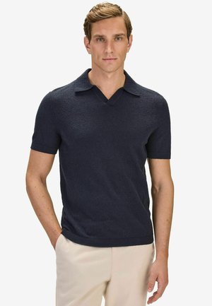 Man med kort blont hår som bär en mörkblå texturerad pikétröja med kort ärm och ljusbeiga byxor, stående med ena handen i fickan.