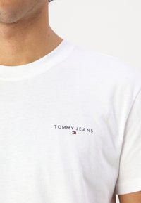 Weißes Baumwoll-T-Shirt mit einem kleinen "TOMMY JEANS"-Logo auf der linken Brust, akzentuiert in rot, weiß und blau.