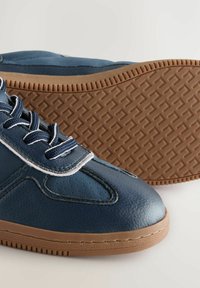 Blå lädersneakers med en slät struktur, som har vita snören, en tanfärgad gummisula och ett geometriskt mönster på yttersulan.