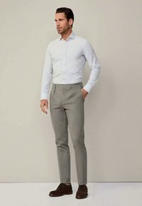 Man standing wearing a light blue striped shirt, gray trousers, and dark brown dress shoes against a plain beige background.
