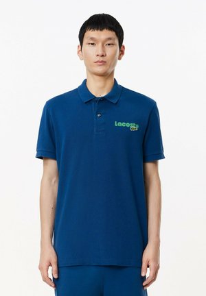 Polo de color azul marino hecho de algodón con cuello plano, tapeta de tres botones y logo verde de Lacoste en el lado izquierdo del pecho. Manga corta.