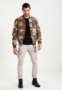 Kurtka bomberka w kamuflażu z naszywkami, ściągaczami na mankietach i w pasie. Noszona z czarnym t-shirtem i beżowymi spodniami cargo, w komplecie z czarnymi butami.