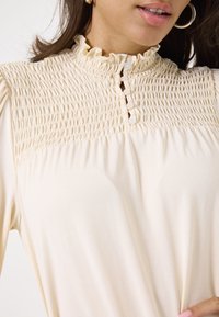 Crème-kleurige blouse met een gesmokte buste, een geschulpte kraag en een knoopsluiting. Zachte stof met een gladde textuur en een vriendelijke val.