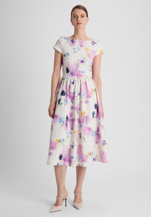 Vrouw draagt een witte midi-jurk met kleurrijke bloemenprints, korte mouwen, boothals en roze puntige hoge hakken, staand tegen een witte achtergrond.