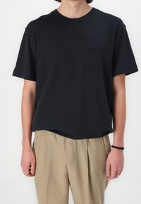 Sort bomuldsskortærmet t-shirt med rund hals og diskret logo på brystet, båret med beige plisserede bukser. Glat tekstur.