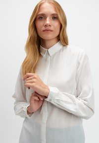 Biała, przezroczysta bluzka z klasycznym kołnierzykiem, długimi rękawami i zapinanymi mankietami z ozdobnymi guzikami. Gładka tekstura z delikatnym połyskiem.