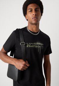 Tricou negru cu un logo verde asemănător vopseli spray "ANTONY MORATO", asortat cu o geantă crossbody texturată neagră și un colier din lanț argintiu.