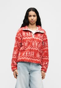 Μπουφάν από φλις με κόκκινο μοτίβο, μισό φερμουάρ στον γιακά και τσέπη, σε συνδυασμό με ανοιχτόχρωμο τζιν με φθορές. Μαλακή υφή και ζωηρό σχέδιο.