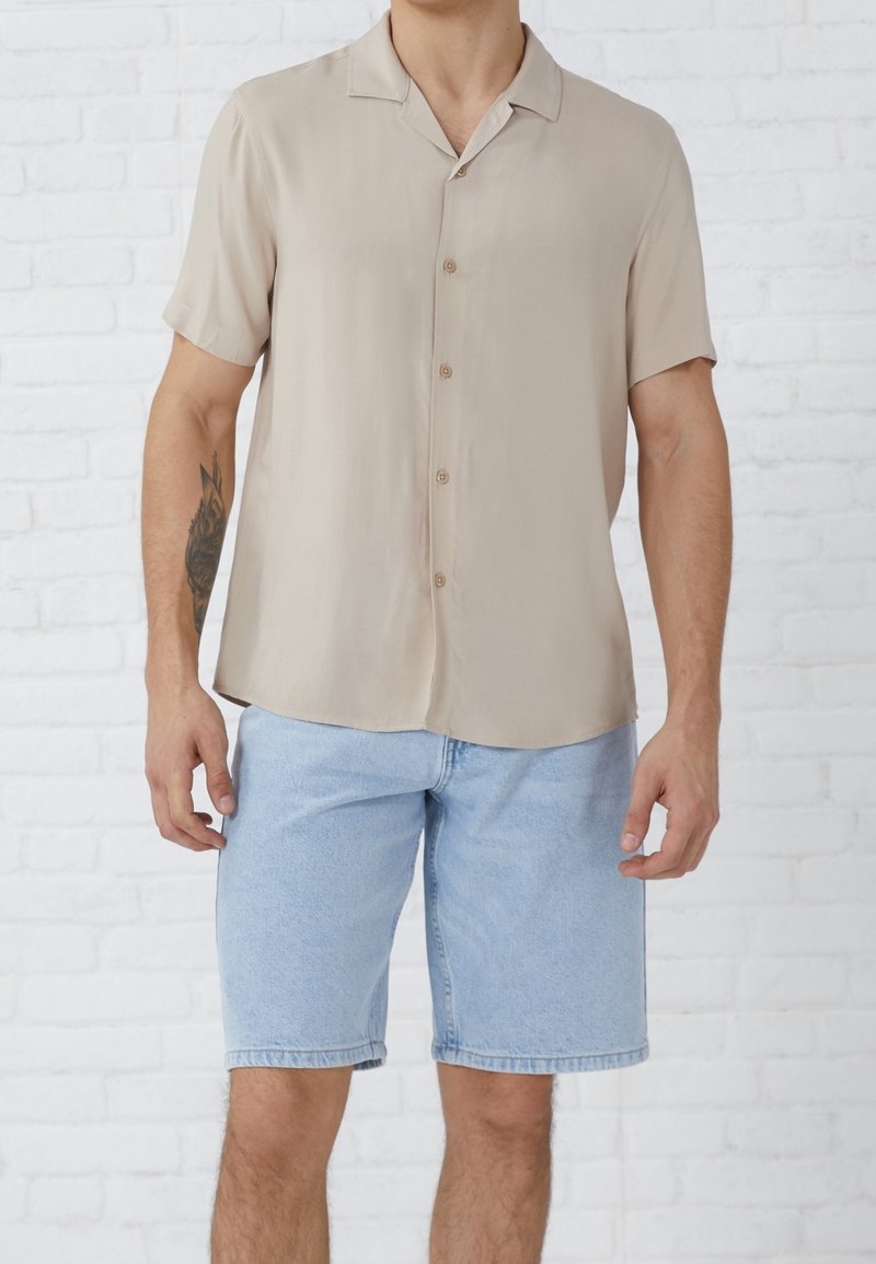 Homme portant une chemise beige à manches courtes boutonnée et un short en jean bleu clair, debout contre un mur en briques blanches.
