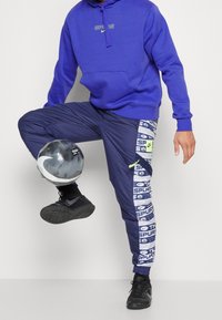 Pantaloni joggers blu con lati decorati e il testo "SPURS", abbinati a un hoodie viola e sneakers nere, mentre tiene in mano un pallone da calcio nero e grigio.