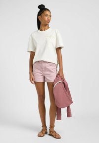 Ljusfärgad bomullströja med ficka och blommönster, tillsammans med rosa denimshorts. Modellen håller en rosa sweatshirt och har på sig bruna sandaler.