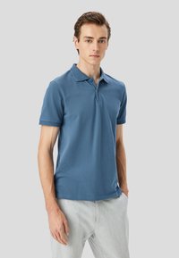 Polo a maniche corte da uomo in blu, con colletto testurizzato e corpo liscio, abbinato a pantaloni grigi chiari.