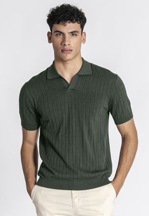 Giovane uomo con capelli ricci che indossa una polo a maniche corte a coste verde scuro e pantaloni beige chiaro, con le mani nelle tasche, rivolto in avanti.