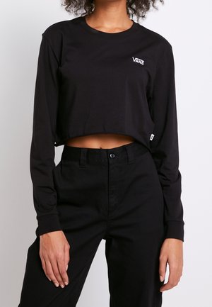 Femme portant un t-shirt Vans noir court à manches longues et un pantalon noir taille haute, debout devant un fond clair uni.