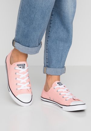 Personne portant un jean bleu clair retroussé et des baskets basses roses Converse All Star avec des lacets blancs, debout sur une surface claire.