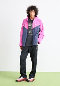Nike farveblokjakke i pink og navy, parret med en sort graphic t-shirt, sorte trackbukser og sneakers med røde accenter på et grønt gulv.