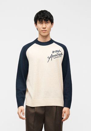 Mladý muž v krémovom a námorníckom svetri s nápisom "Hotel Amour" na hrudi, v hnedých nohaviciach, stojaci pred jednoduchým pozadím.