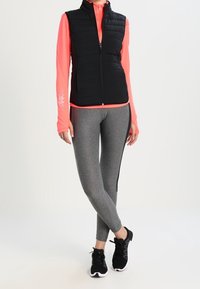 Svart pufferväst över en rosa långärmad topp, tillsammans med grå leggings med svarta sidostrecken, och svarta träningsskor.